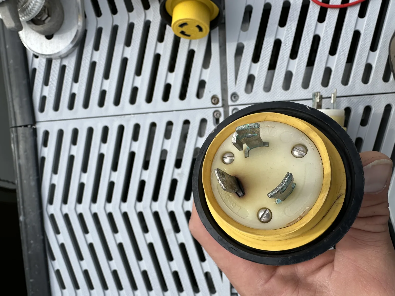 A corroded, heat-discolored shore-power plug held up against a boat's electrical panel, its prongs worn from years of dockside use